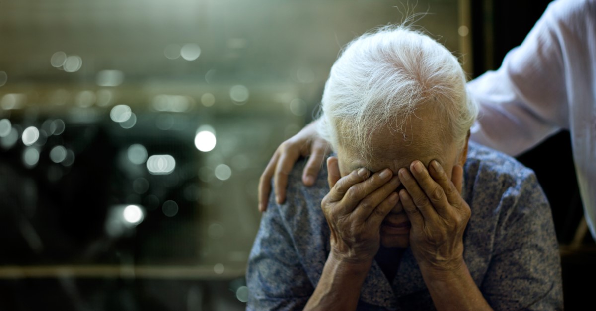 Staršia žena má zdravotný problém, hlavu má podopretú rukami a za ramenu ju drží osoba