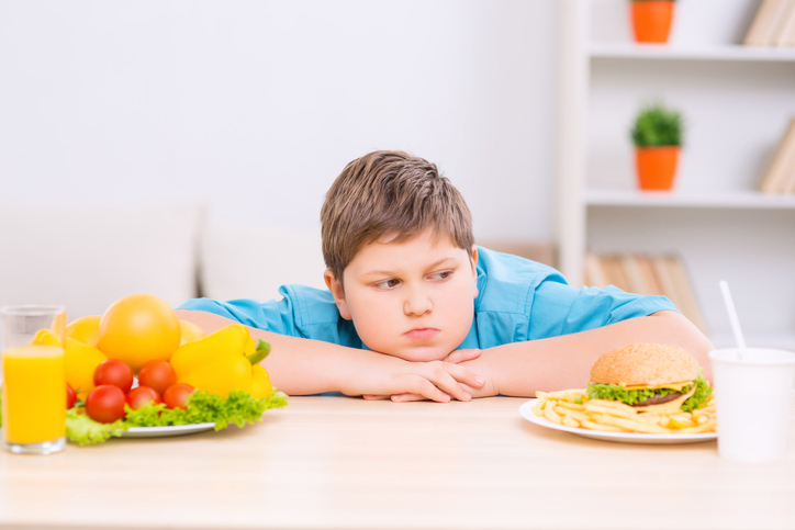 Tučný chlapec medzi dvoma tanierami so zdravým a nezdravým jedlom.
