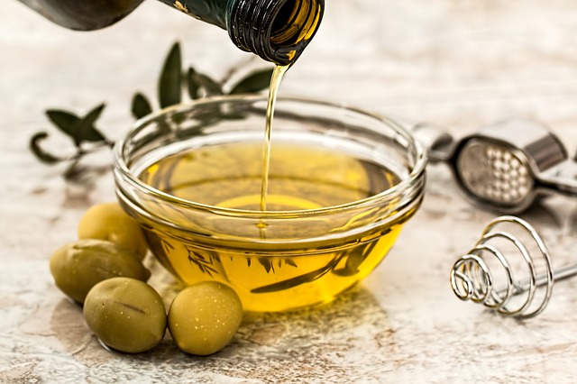 Do sklenenej misky sa nalieva olivový olej zo sklenenej fľaše. Okolo misky sú uložené zelené olivy. 